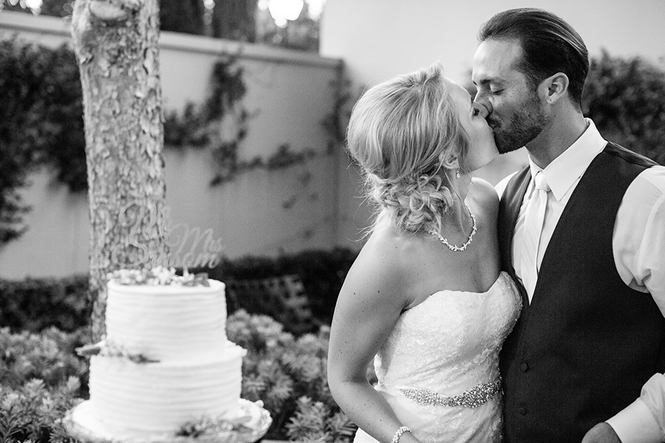 Orange county outdoor wedding at the oak canyon nature center bride form fitting mermaid style strapless gown with a crystal belt and sweetheart neckline with a long veil and groom slate blue notch lapel suit with a matching vest and white dress shirt with a long white tie and white floral boutonniere kissing after cutting cake black and white photo