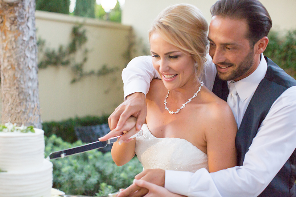 Orange county outdoor wedding at the oak canyon nature center bride form fitting mermaid style strapless gown with a crystal belt and sweetheart neckline with a long veil and groom slate blue notch lapel suit with a matching vest and white dress shirt with a long white tie and white floral boutonniere cutting cake