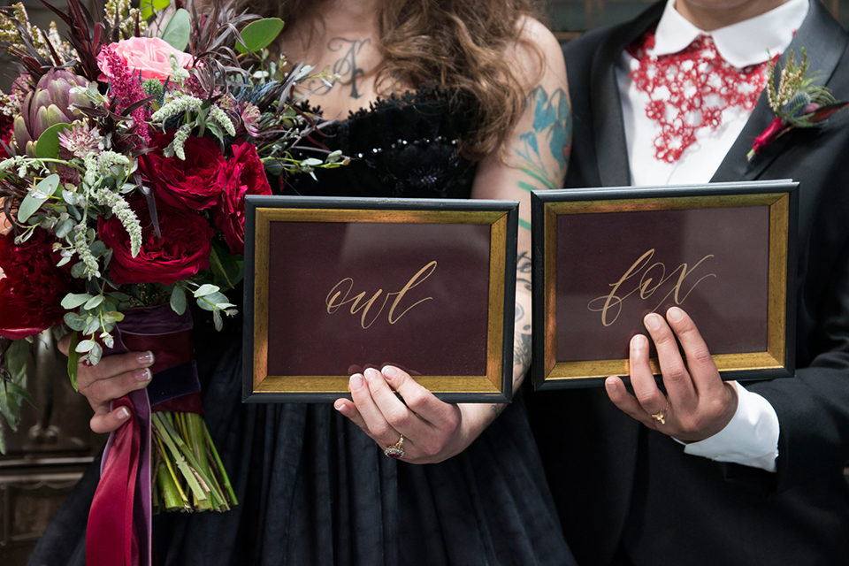 Fox and the owl wedding shot at big daddys antiques bride black womens shawl lapel tuxedo with a white dress shirt and red lace design on neckline with red floral boutonniere with bride black ball gown with thin straps and sweetheart neckline close up holding signs and bride holding red floral bridal bouquet