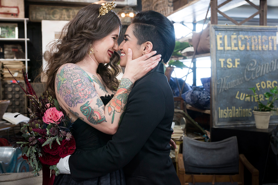 Fox and the owl wedding shot at big daddys antiques bride black womens shawl lapel tuxedo with a white dress shirt and red lace design on neckline with red floral boutonniere with bride black ball gown with thin straps and sweetheart neckline hugging close up and bride holding red floral bridal bouquet