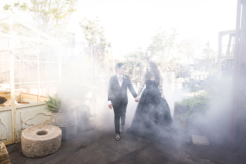 Fox and the owl wedding shot at big daddys antiques bride black womens shawl lapel tuxedo with a white dress shirt and red lace design on neckline with red floral boutonniere with bride black ball gown with thin straps and sweetheart neckline holding hands