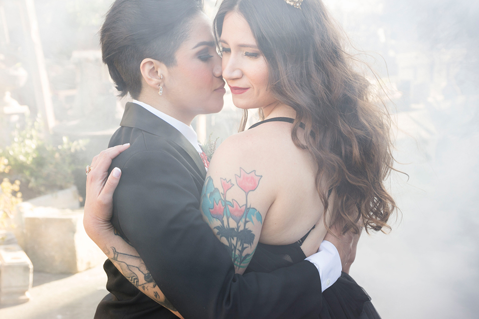 Fox and the owl wedding shot at big daddys antiques bride black womens shawl lapel tuxedo with a white dress shirt and red lace design on neckline with red floral boutonniere with bride black ball gown with thin straps and sweetheart neckline hugging close up