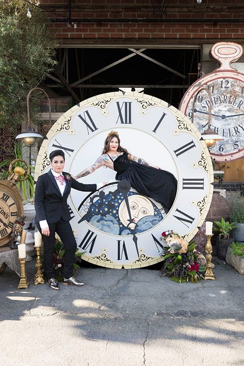 Fox and the owl wedding shot at big daddys antiques bride black womens shawl lapel tuxedo with a white dress shirt and red lace design on neckline with red floral boutonniere with bride black ball gown with thin straps and sweetheart neckline ceremony holding hands in clock