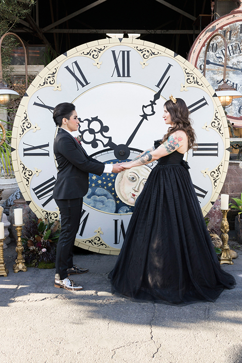 Fox and the owl wedding shot at big daddys antiques bride black womens shawl lapel tuxedo with a white dress shirt and red lace design on neckline with red floral boutonniere with bride black ball gown with thin straps and sweetheart neckline ceremony standing holding hands