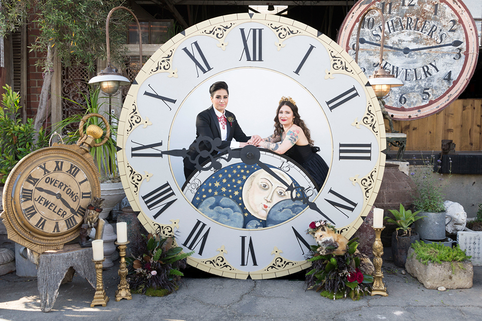 Fox and the owl wedding shot at big daddys antiques bride black womens shawl lapel tuxedo with a white dress shirt and red lace design on neckline with red floral boutonniere with bride black ball gown with thin straps and sweetheart neckline sitting in clock ceremony