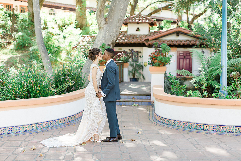 Rancho las lomas outdoor wedding shoot with spanish inspiration bride form fitting lace gown with beaded detail and a plunging neckline with open back design with groom slate blue notch lapel suit with a matching vest and white dress shirt with a matching slate pipe edge bow tie holding hands and kissing