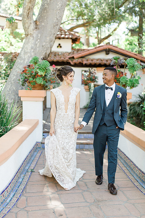 Rancho las lomas outdoor wedding shoot with spanish inspiration bride form fitting lace gown with beaded detail and a plunging neckline with open back design with groom slate blue notch lapel suit with a matching vest and white dress shirt with a matching slate pipe edge bow tie holding hands and walking