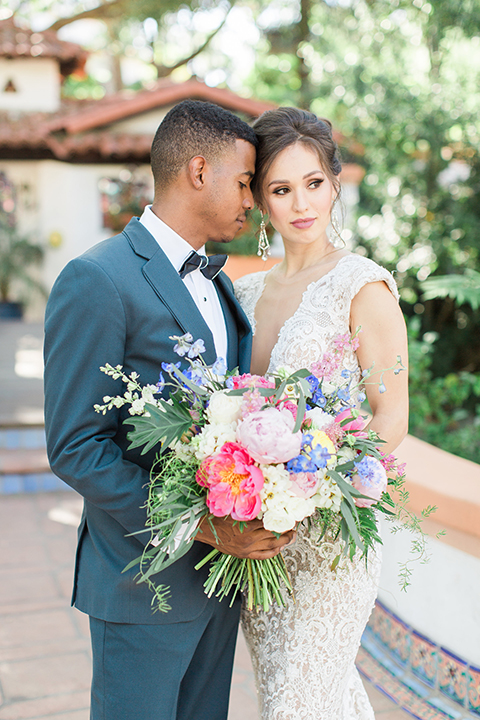 Rancho las lomas outdoor wedding shoot with spanish inspiration bride form fitting lace gown with beaded detail and a plunging neckline with open back design with groom slate blue notch lapel suit with a matching vest and white dress shirt with a matching slate pipe edge bow tie hugging close up and bride holding pink and blue colorful floral bridal bouquet