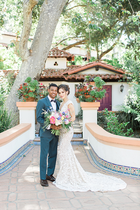 Rancho las lomas outdoor wedding shoot with spanish inspiration bride form fitting lace gown with beaded detail and a plunging neckline with open back design with groom slate blue notch lapel suit with a matching vest and white dress shirt with a matching slate pipe edge bow tie standing and hugging bride holding pink and blue floral bridal bouquet