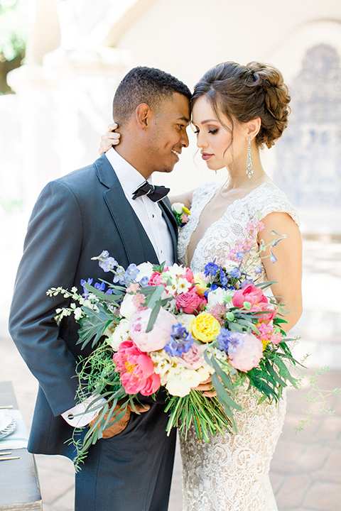 Rancho las lomas outdoor wedding shoot with spanish inspiration bride form fitting lace gown with beaded detail and a plunging neckline with open back design with groom slate blue notch lapel suit with a matching vest and white dress shirt with a matching slate pipe edge bow tie hugging close up and bride holding pink and blue colorful bridal bouquet