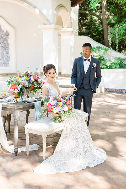 Rancho las lomas outdoor wedding shoot with spanish inspiration bride form fitting lace gown with beaded detail and a plunging neckline with open back design with groom slate blue notch lapel suit with a matching vest and white dress shirt with a matching slate pipe edge bow tie sitting at table bride sitting on bench with pink and blue colorful floral bridal bouquet