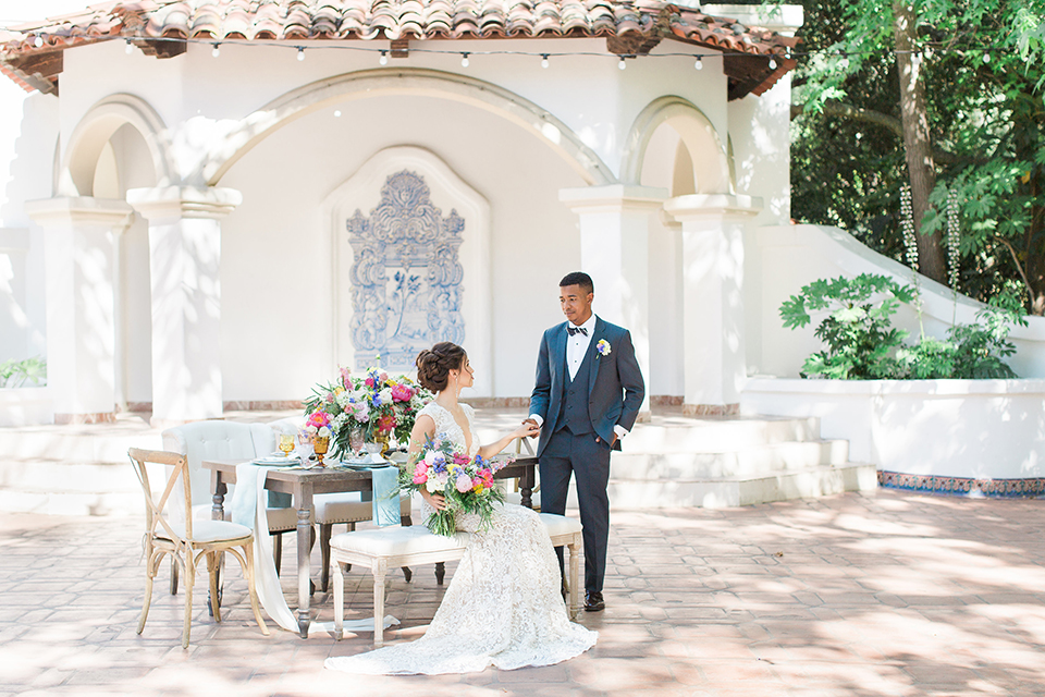 Rancho las lomas outdoor wedding shoot with spanish inspiration bride form fitting lace gown with beaded detail and a plunging neckline with open back design with groom slate blue notch lapel suit with a matching vest and white dress shirt with a matching slate pipe edge bow tie bride sitting at table with pink and blue colorful floral bridal bouquet