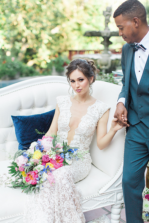 Rancho las lomas outdoor wedding shoot with spanish inspiration bride form fitting lace gown with beaded detail and a plunging neckline with open back design with groom slate blue notch lapel suit with a matching vest and white dress shirt with a matching slate pipe edge bow tie bride sitting on white couch holding hands and holding pink and blue colorful floral bridal bouquet