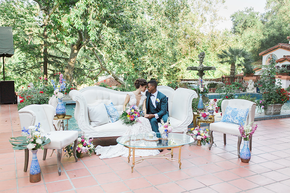 Rancho las lomas outdoor wedding shoot with spanish inspiration bride form fitting lace gown with beaded detail and a plunging neckline with open back design with groom slate blue notch lapel suit with a matching vest and white dress shirt with a matching slate pipe edge bow tie sitting on white couch with bride holding pink and blue floral bridal bouquet