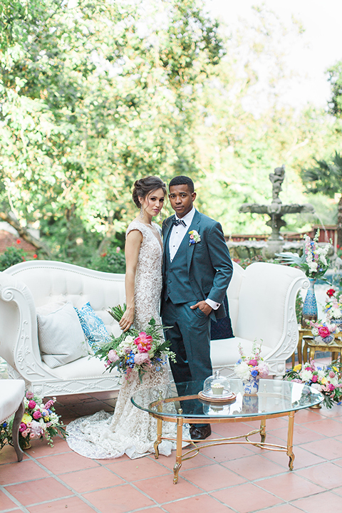 Rancho las lomas outdoor wedding shoot with spanish inspiration bride form fitting lace gown with beaded detail and a plunging neckline with open back design with groom slate blue notch lapel suit with a matching vest and white dress shirt with a matching slate pipe edge bow tie standing by couch and bride holding pink and blue colorful bridal floral bouquet