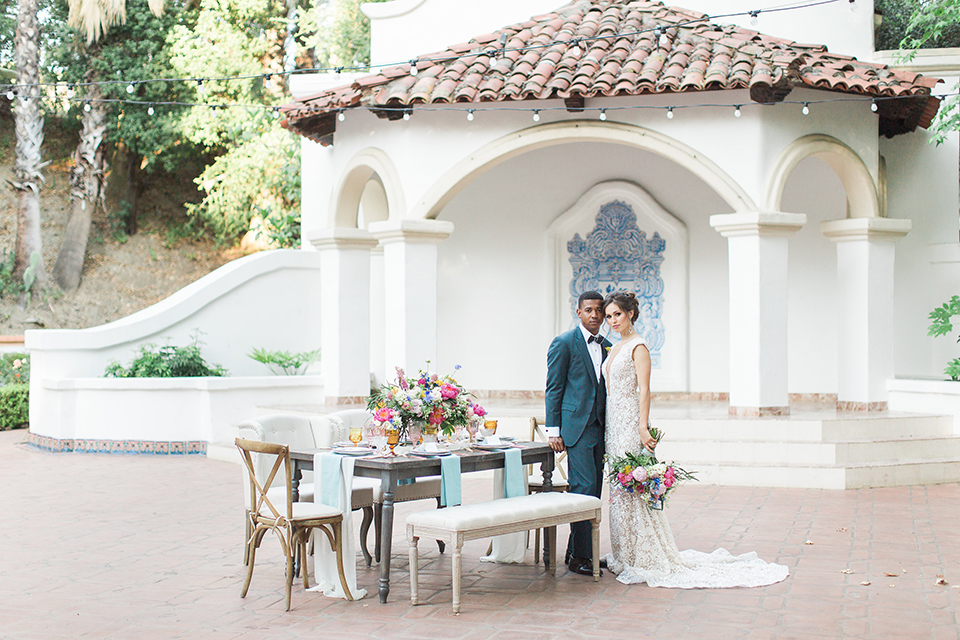 Rancho las lomas outdoor wedding shoot with spanish inspiration bride form fitting lace gown with beaded detail and a plunging neckline with open back design with groom slate blue notch lapel suit with a matching vest and white dress shirt with a matching slate pipe edge bow tie standing by table bride holding pink and blue colorful floral bridal bouquet