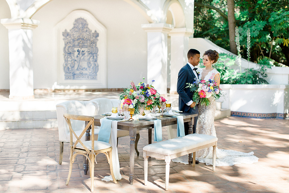 Rancho las lomas outdoor wedding shoot with spanish inspiration bride form fitting lace gown with beaded detail and a plunging neckline with open back design with groom slate blue notch lapel suit with a matching vest and white dress shirt with a matching slate pipe edge bow tie standing by table bride holding pink and blue colorful floral bridal bouquet