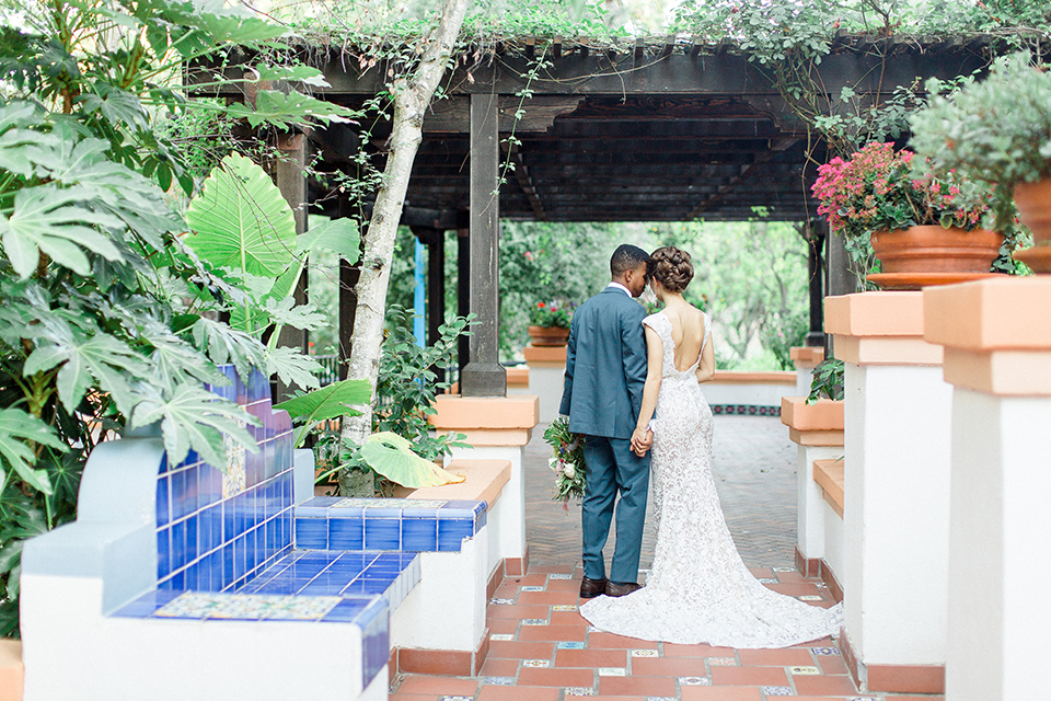 Rancho las lomas outdoor wedding shoot with spanish inspiration bride form fitting lace gown with beaded detail and a plunging neckline with open back design with groom slate blue notch lapel suit with a matching vest and white dress shirt with a matching slate pipe edge bow tie holding hands and groom holding pink and blue colorful floral bridal bouquet