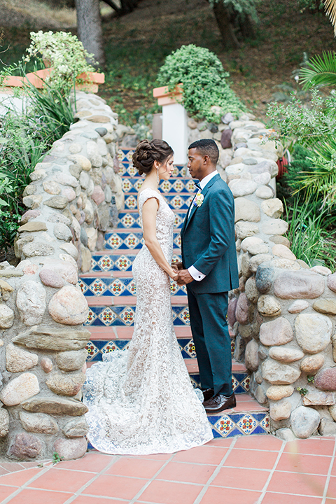 Rancho las lomas outdoor wedding shoot with spanish inspiration bride form fitting lace gown with beaded detail and a plunging neckline with open back design with groom slate blue notch lapel suit with a matching vest and white dress shirt with a matching slate pipe edge bow tie holding hands and stairs