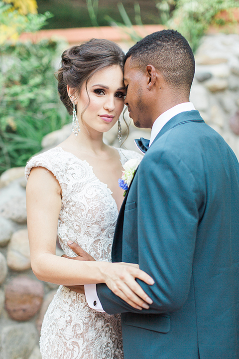 Rancho las lomas outdoor wedding shoot with spanish inspiration bride form fitting lace gown with beaded detail and a plunging neckline with open back design with groom slate blue notch lapel suit with a matching vest and white dress shirt with a matching slate pipe edge bow tie holding hands and hugging close up