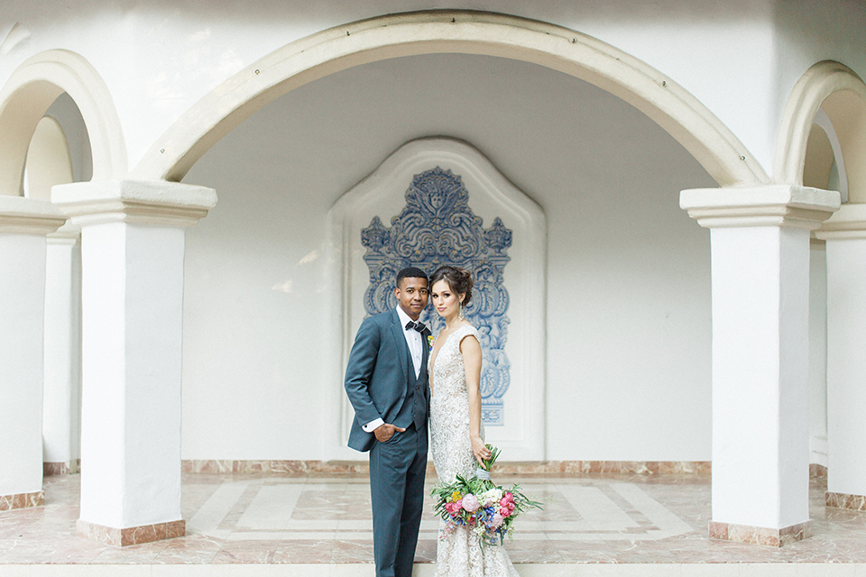 Rancho las lomas outdoor wedding shoot with spanish inspiration bride form fitting lace gown with beaded detail and a plunging neckline with open back design with groom slate blue notch lapel suit with a matching vest and white dress shirt with a matching slate pipe edge bow tie hugging and bride holding pink and blue colorful floral bridal bouquet