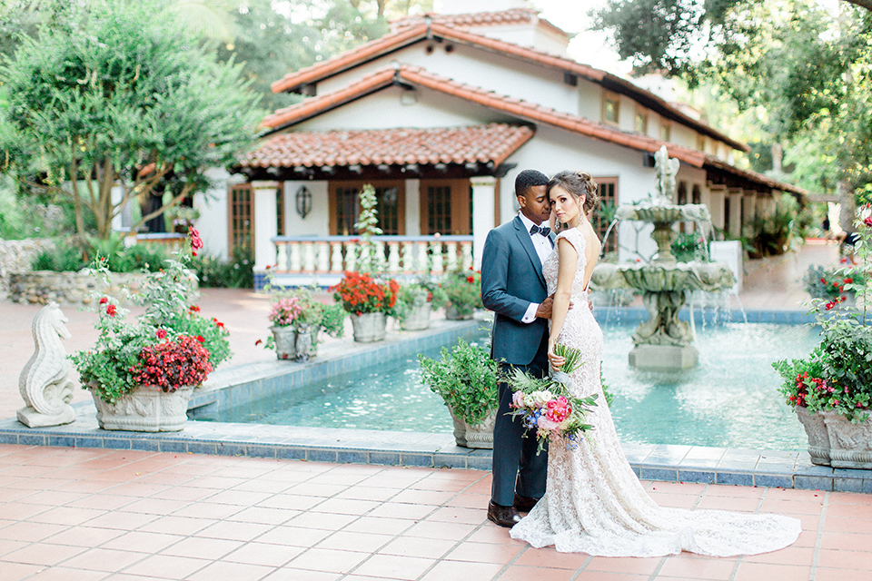 Rancho las lomas outdoor wedding shoot with spanish inspiration bride form fitting lace gown with beaded detail and a plunging neckline with open back design with groom slate blue notch lapel suit with a matching vest and white dress shirt with a matching slate pipe edge bow tie hugging and bride holding pink and blue colorful floral bridal bouquet