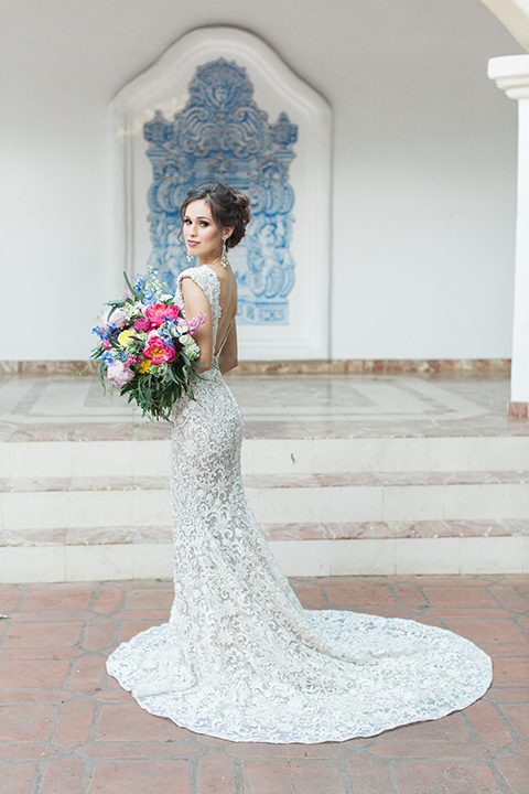 Rancho las lomas outdoor wedding shoot with spanish inspiration bride form fitting lace gown with beaded detail and a plunging neckline with open back design holding pink and blue colorful floral bridal bouquet