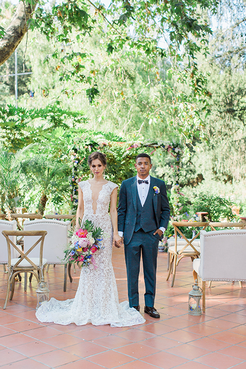 Rancho las lomas outdoor wedding shoot with spanish inspiration bride form fitting lace gown with beaded detail and a plunging neckline with open back design with groom slate blue notch lapel suit with a matching vest and white dress shirt with a matching slate pipe edge bow tie holding hands and bride holding pink and blue colorful floral bridal bouquet