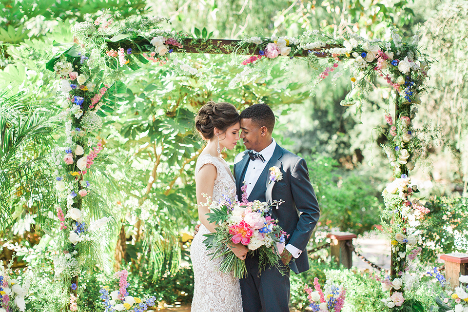 Rancho las lomas outdoor wedding shoot with spanish inspiration bride form fitting lace gown with beaded detail and a plunging neckline with open back design with groom slate blue notch lapel suit with a matching vest and white dress shirt with a matching slate pipe edge bow tie hugging and bride holding pink and blue colorful floral bridal bouquet