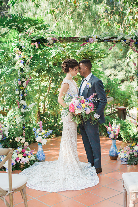 Rancho las lomas outdoor wedding shoot with spanish inspiration bride form fitting lace gown with beaded detail and a plunging neckline with open back design with groom slate blue notch lapel suit with a matching vest and white dress shirt with a matching slate pipe edge bow tie hugging and bride holding pink and blue colorful floral bridal bouquet