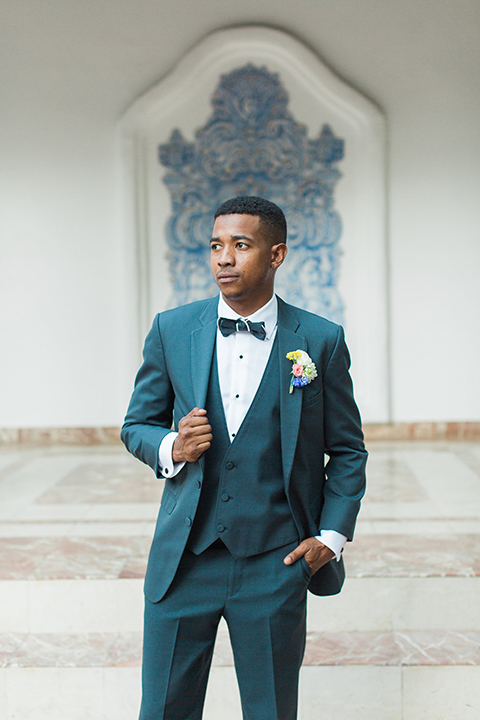 Rancho las lomas outdoor wedding with spanish inspiration groom slate blue notch lapel suit with a matching vest and white dress shirt with a matching slate blue pipe edge bow tie and white floral boutonniere holding jacket