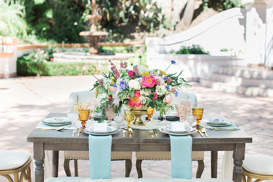 Rancho las lomas outdoor wedding with spanish inspiration table set up light grey wood table with white vintage chairs and light blue napkin decor with white and blue place settings with pink and blue colorful flower centerpiece decor and yellow glasses with gold silverware