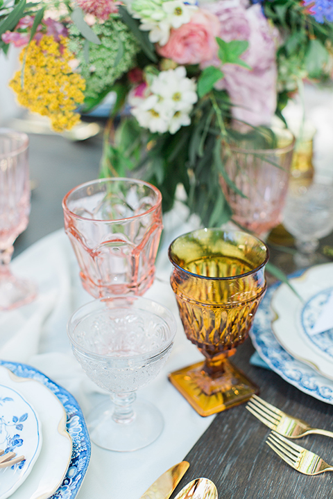 Rancho las lomas outdoor wedding with spanish inspiration table set up light grey wood table with white vintage chairs and light blue napkin decor with white and blue place settings with pink and blue colorful flower centerpiece decor and yellow glasses with gold silverware