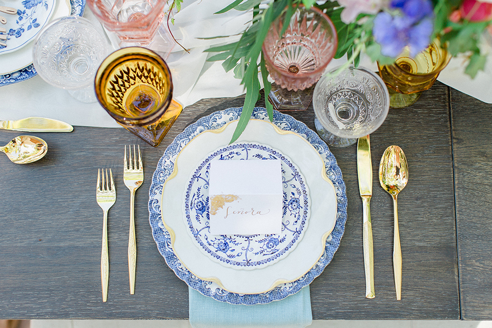 Rancho las lomas outdoor wedding with spanish inspiration table set up light grey wood table with white vintage chairs and light blue napkin decor with white and blue place settings with pink and blue colorful flower centerpiece decor and yellow glasses with gold silverware