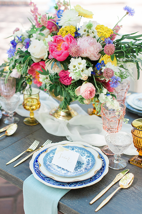 Rancho las lomas outdoor wedding with spanish inspiration table set up light grey wood table with white vintage chairs and light blue napkin decor with white and blue place settings with pink and blue colorful flower centerpiece decor and yellow glasses with gold silverware