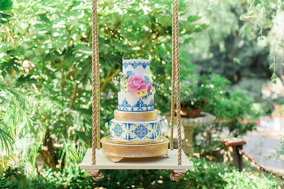 Rancho las lomas outdoor wedding with spanish inspiration table set up light grey wood table with white vintage chairs and light blue napkin decor with white and blue place settings with pink and blue colorful flower centerpiece decor and yellow glasses with gold silverware with wedding cake three tier white and blue patterned cake with gold accents and pink flowers hanging on wood table