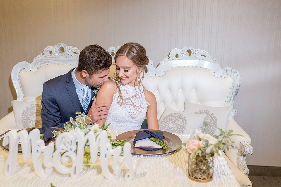 San diego wedding at the hilton bayside bride form fitting lace gown with a high halter neckline and slit in leg area with groom navy notch lapel suit with a light blue dress shirt and a light blue striped long tie with a white and green floral boutonniere sitting at table