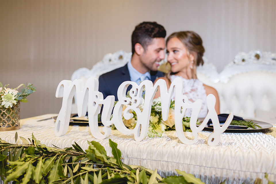 San diego wedding at the hilton bayside bride form fitting lace gown with a high halter neckline and slit in leg area with groom navy notch lapel suit with a light blue dress shirt and a light blue striped long tie with a white and green floral boutonniere sitting at reception table smiling