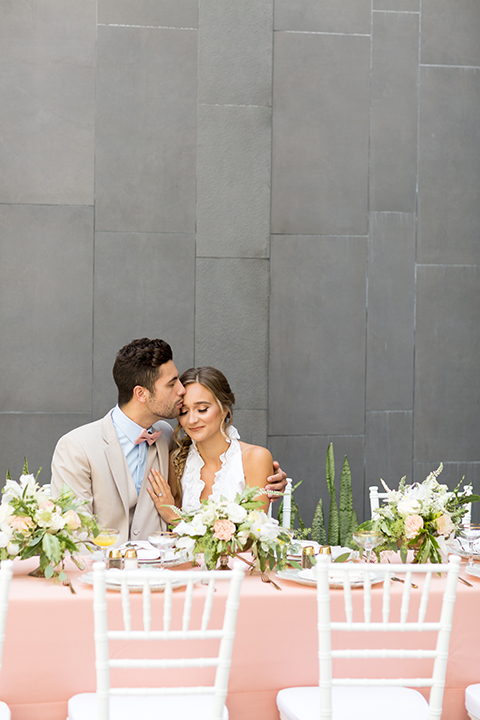 San diego wedding at the hilton bayside bride short white dress with a lace halter bodice and chiffon skirt with groom tan notch lapel suit with a light blue dress shirt and a blush pink bow tie sitting at table kissing