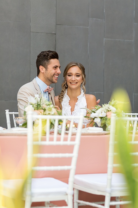 San diego wedding at the hilton bayside bride short white dress with a lace halter bodice and chiffon skirt with groom tan notch lapel suit with a light blue dress shirt and a blush pink bow tie sitting at table smiling