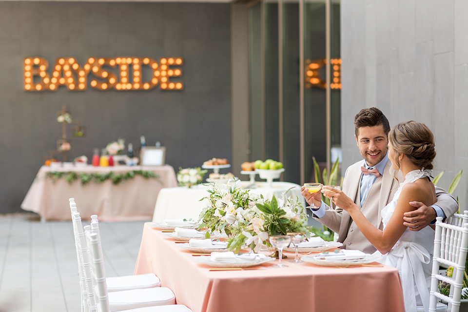 San diego wedding at the hilton bayside bride short white dress with a lace halter bodice and chiffon skirt with groom tan notch lapel suit with a light blue dress shirt and a blush pink bow tie sitting at table smiling and holding drinks