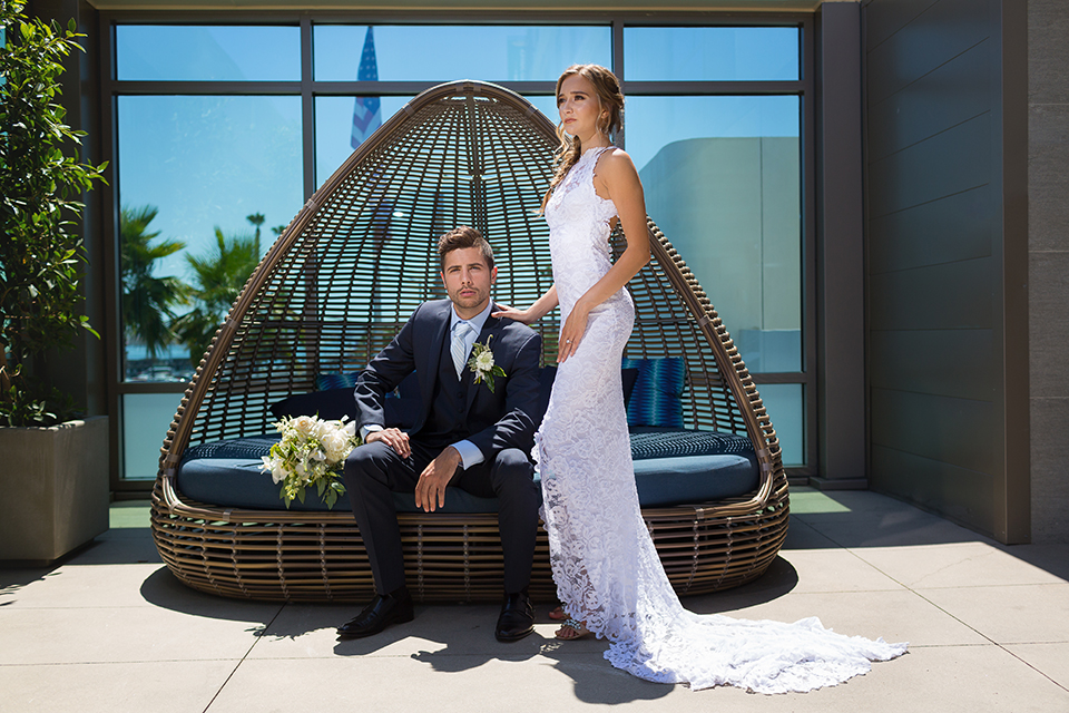 San diego wedding at the hilton bayside bride form fitting lace gown with a high halter neckline and slit in leg area with groom navy notch lapel suit with a light blue dress shirt and a light blue striped long tie with a white and green floral boutonniere sitting on couch with green and white floral bridal bouquet with bride standing