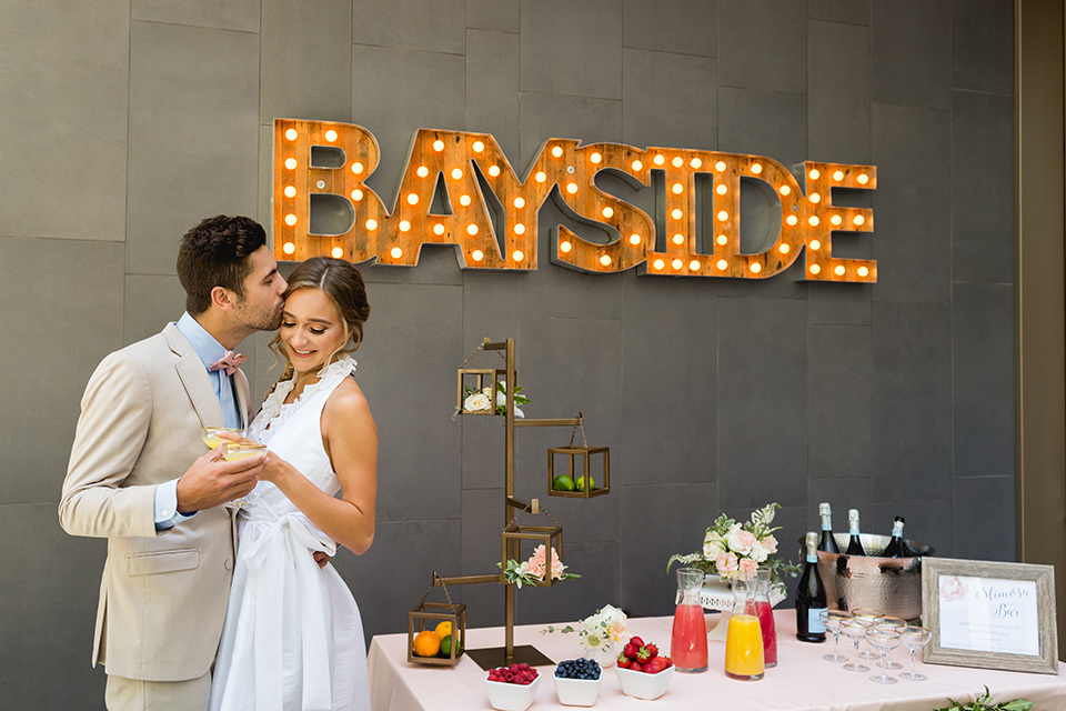 San diego wedding at the hilton bayside bride short white dress with a lace halter bodice and chiffon skirt with groom tan notch lapel suit with a light blue dress shirt and a blush pink bow tie standing by drinks table hugging and holding drinks with sign in background