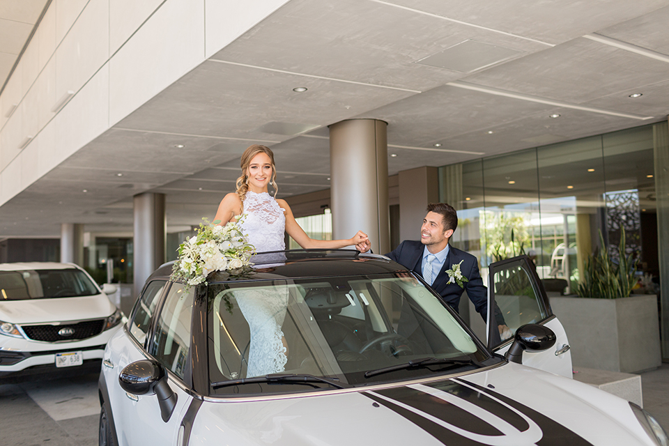 San diego wedding at the hilton bayside bride form fitting lace gown with a high halter neckline and slit in leg area with groom navy notch lapel suit with a light blue dress shirt and a light blue striped long tie with a white and green floral boutonniere standing in car with bride holding white and green floral bridal bouquet