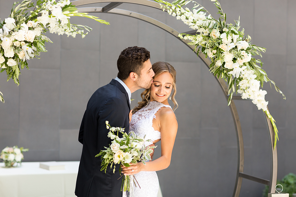 San diego wedding at the hilton bayside bride form fitting lace gown with a high halter neckline and slit in leg area with groom navy notch lapel suit with a light blue dress shirt and a light blue striped long tie with a white and green floral boutonniere standing and hugging and kissing in ceremony bride holding white and green floral bridal bouquet