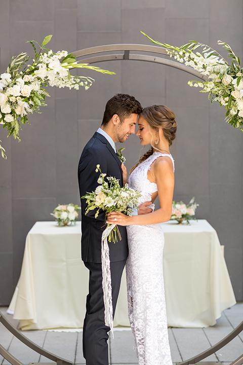 San diego wedding at the hilton bayside bride form fitting lace gown with a high halter neckline and slit in leg area with groom navy notch lapel suit with a light blue dress shirt and a light blue striped long tie with a white and green floral boutonniere standing and hugging during ceremony bride holding white and green floral bridal bouquet with white ribbon decor