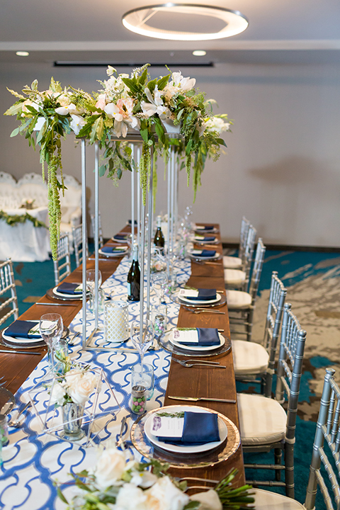 San diego wedding at the hilton bayside table set up brown wood table with white and clear place settings with blue linen napkin decor and white and blue table runner with white and green tall flower centerpiece decor in clear vases with white chiavari chairs