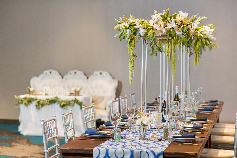 San diego wedding at the hilton bayside table set up brown wood table with white and clear place settings with blue linen napkin decor and white and blue table runner with white and green tall flower centerpiece decor in clear vases with white chiavari chairs