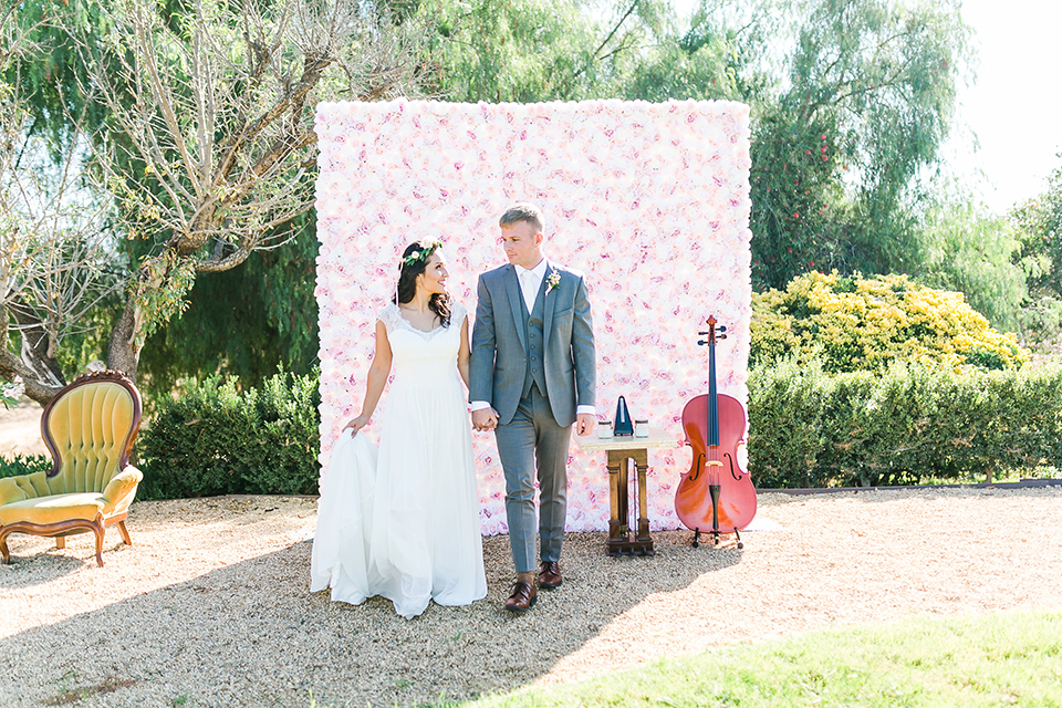 Temecula outdoor wedding at bel vino winery bride a line chiffon gown with straps and lace detail on bodice with a sweetheart neckline and white and green flower crown with groom grey notch lapel suit with a matching vest and white dress shirt with a long white tie and white and pink floral boutonniere holding hands