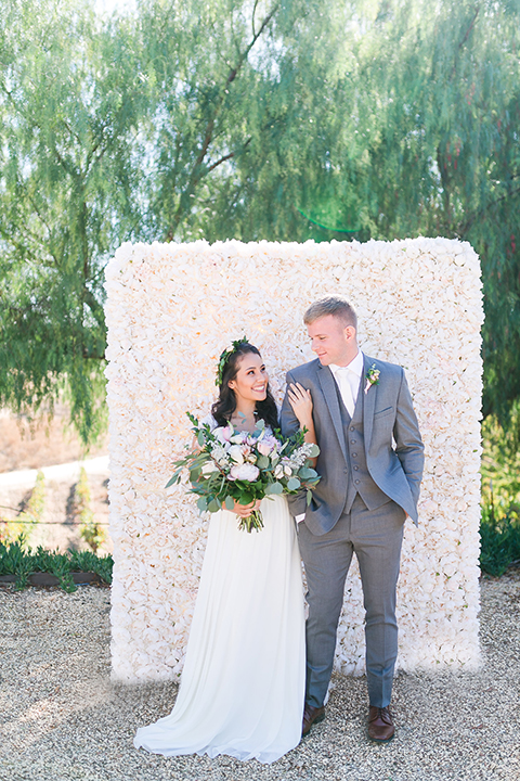 Temecula outdoor wedding at bel vino winery bride a line chiffon gown with straps and lace detail on bodice with a sweetheart neckline and white and green flower crown with groom grey notch lapel suit with a matching vest and white dress shirt with a long white tie and white and pink floral boutonniere holding hands